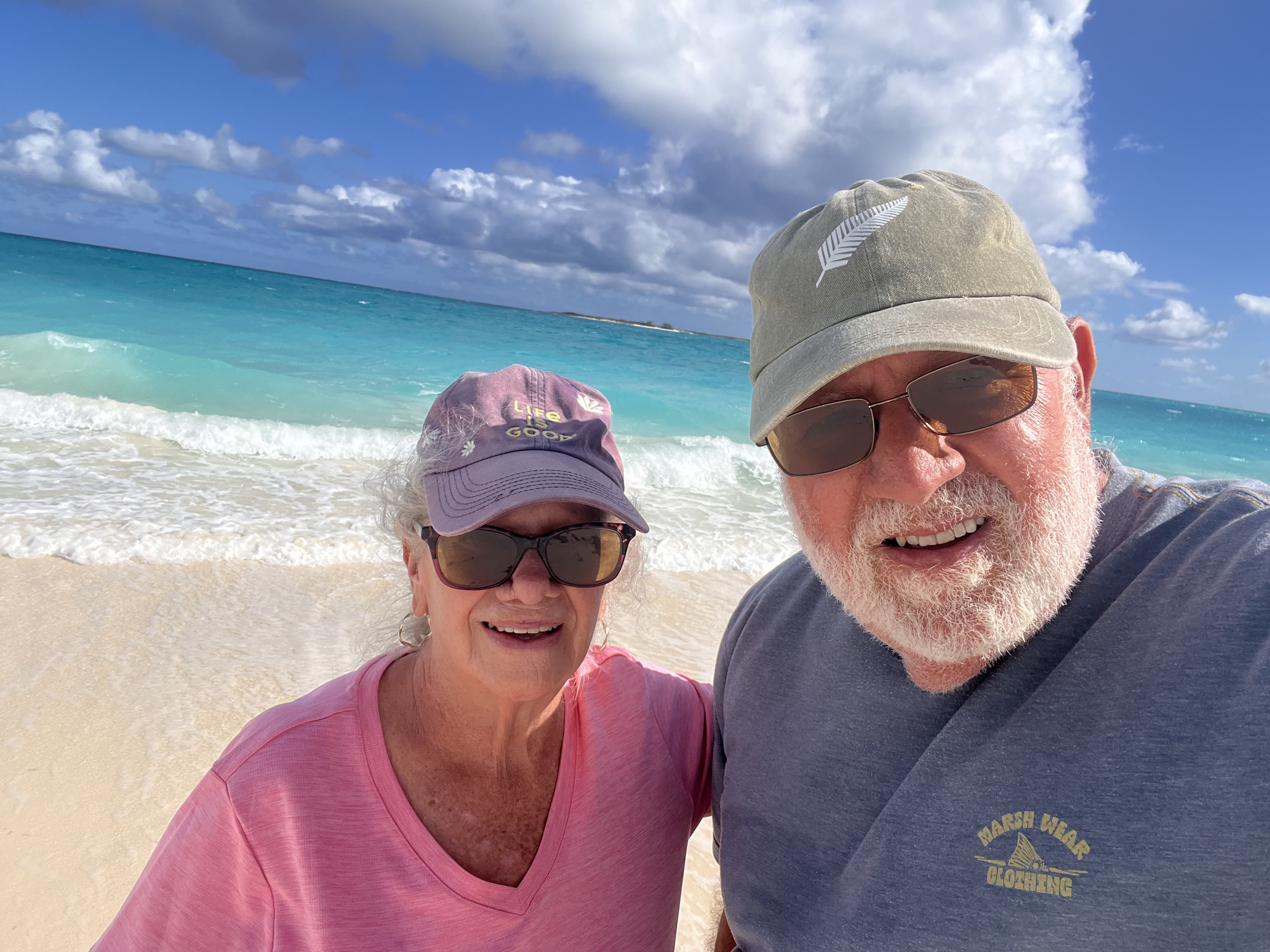 Two adults smiling together on a bright beach with turquoise water behind them.