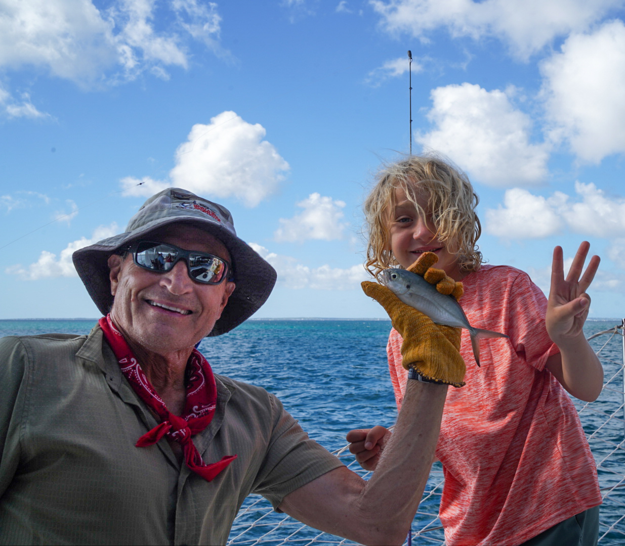 An adult and child smiling aboard a sailboat, holding a small fish above the sea.